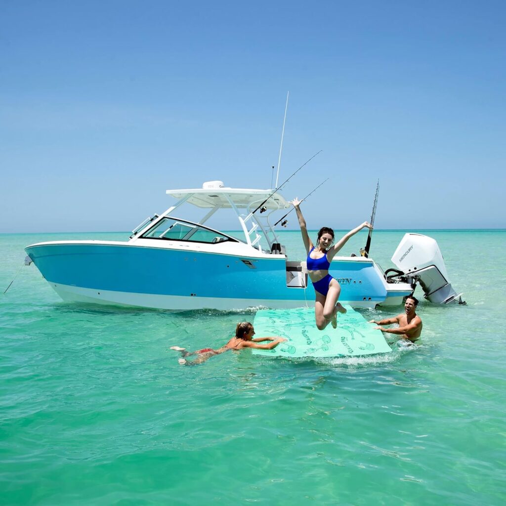 An Aqua Lily Pad floating mat adjacent to a small boat. Bathers are holding onto the mat while a woman jumps off of the mat into the water.